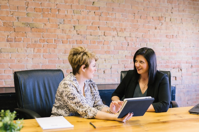 Two professionals seated at a wooden table in a modern office with exposed brick walls engage in a discussion about HSA email templates. One person holds a tablet, explaining details while the other listens attentively.