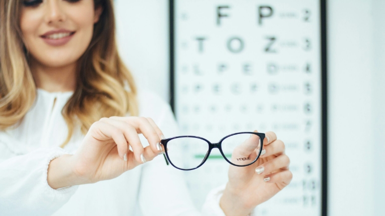 Glasses held in front of an eye chart by a smiling woman, symbolizing eye care affordability through FSA Options.
