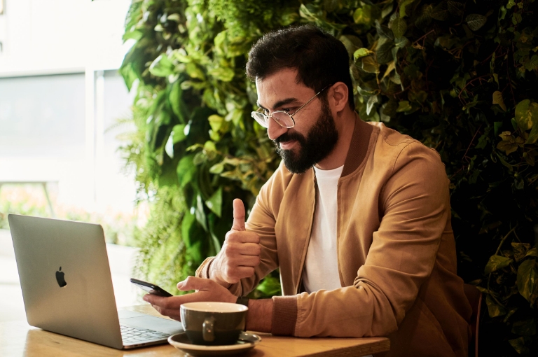 A professional seated at a wooden table uses a smartphone and laptop while giving a thumbs-up, symbolizing satisfaction with HSA flyers. Surrounded by lush green plants and natural light, the workspace includes a coffee cup and modern tech setup.