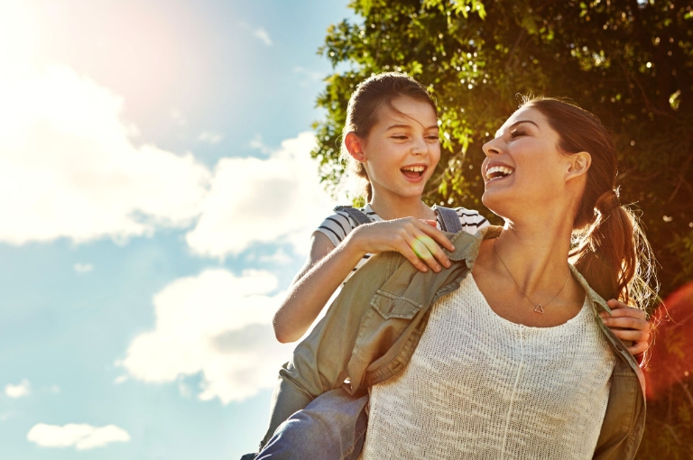 Mother giving daughter a piggyback ride outdoors, symbolizing family health and savings benefits supported by HSA communication toolkits.