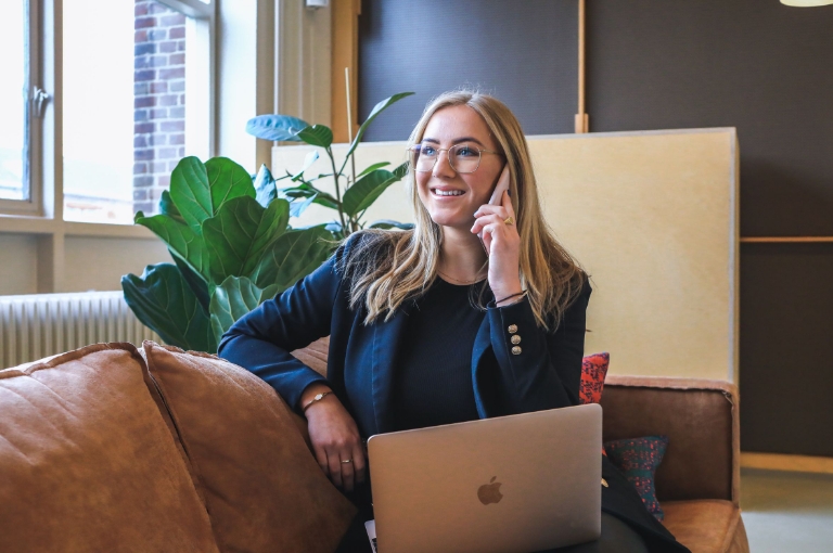 A professional seated on a brown couch in a modern, well-lit office uses a smartphone and laptop, representing engagement with HSA online resources. The individual wears a dark blazer and glasses, with a laptop open on their lap. Large windows, green plants, and contemporary decor emphasize a productive and accessible digital workspace.