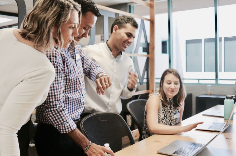 Four colleagues collaborate around a desk in a modern office, with one person pointing at a laptop screen and another checking a watch. The scene reflects a productive team environment ideal for introducing HSA getting started resources.