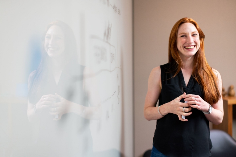 A professional stands in front of a whiteboard with faint diagrams and writing, smiling and appearing engaged in a presentation. The setting suggests a workplace discussion using visual aids, ideal for showcasing HSA slide deck resources that support effective employee communication and benefits education.