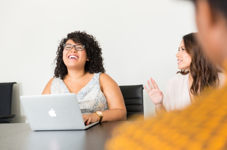 Three professionals collaborate around a conference table in a bright, modern office, with one person smiling while using a laptop and another gesturing during discussion. This engaging workplace scene reflects the accessibility of HSA video resources and digital communication tools for employee benefits.